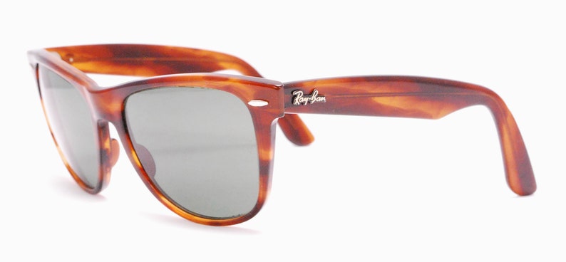 Puede incluir: Gafas de sol Ray-Ban Wayfarer de carey con lentes verdes. Las gafas tienen un logotipo Ray-Ban plateado en la patilla derecha.