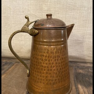 Puede incluir: Una jarra de cobre martillado con asa y tapa de latón. La jarra tiene una superficie texturizada y una base redondeada.