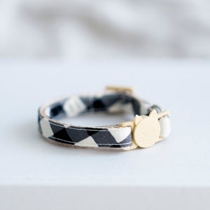 May include: A black and white patterned cat collar with a gold-colored cat-shaped charm. The collar is resting on a white book titled "THE KINFOLK HOME". The background is a soft, blurred white.
