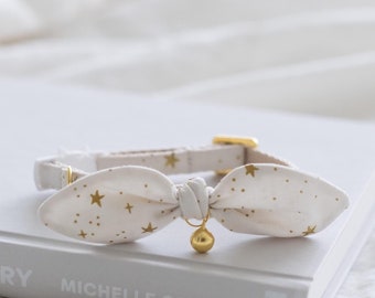 Puede incluir: Un collar para gato color crema con diseño de lazo, adornado con estrellas doradas. El collar tiene una hebilla dorada y una pequeña campana dorada. Descansa sobre un libro gris con el título "Light We Carry".
