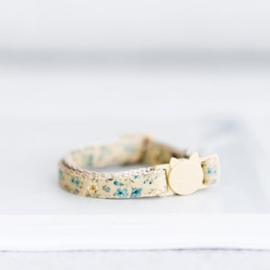 May include: A beige cat collar with a floral pattern of small blue flowers. The collar has a cream-colored cat-shaped buckle. The collar is approximately 0.5 inches wide.