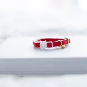 May include: A red cat collar with a white cat-shaped buckle and a small gold-colored bell. The collar is made of a smooth material and is resting on a white surface. The collar is designed for a pet.