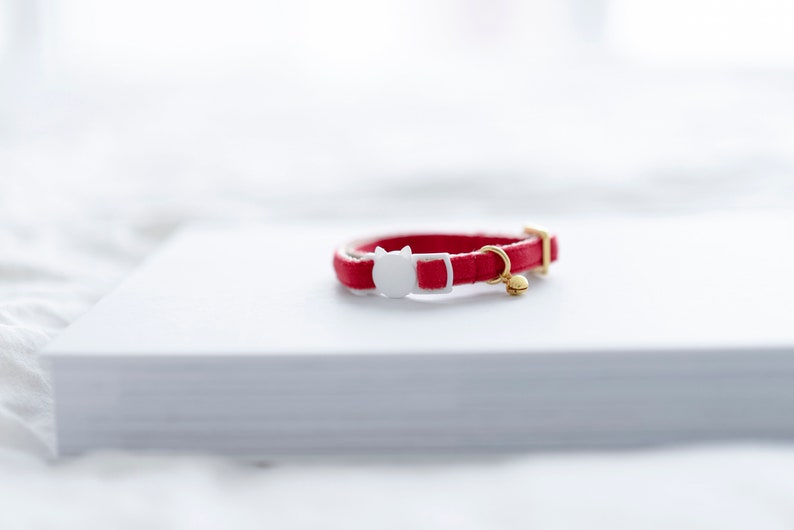 May include: A red cat collar with a white cat-shaped buckle and a small gold-colored bell. The collar is made of a smooth material and is resting on a white surface. The collar is designed for a pet.
