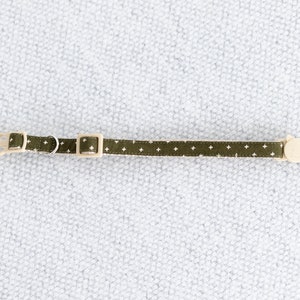 May include: A green cat collar with a white cross pattern. The collar has a beige plastic buckle and a small metal ring. A cat-shaped charm is attached to the end. The collar measures approximately 12 inches in length.