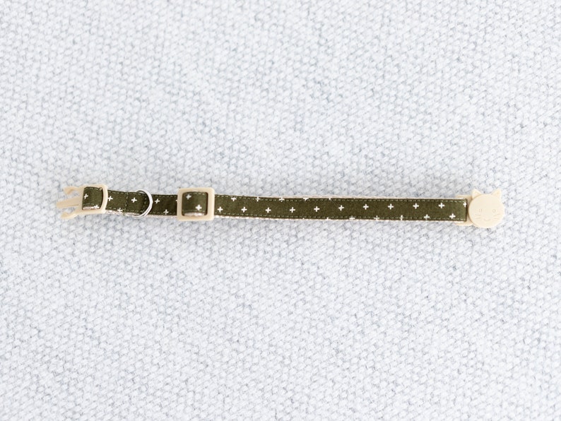May include: A green cat collar with a white cross pattern. The collar has a beige plastic buckle and a small metal ring. A cat-shaped charm is attached to the end. The collar measures approximately 12 inches in length.