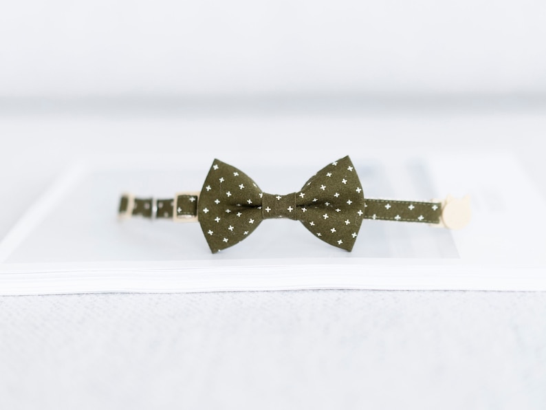 May include: An olive green cat collar with a matching bow tie. The bow tie and collar feature a white star pattern. The collar has a gold-colored buckle and a small, beige cat-shaped charm.