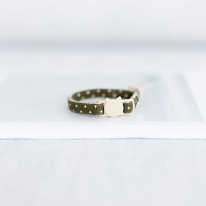 May include: Olive green cat collar with white plus signs and small white dots. The collar has a cream-colored cat-shaped buckle. A stylish accessory for your feline friend.
