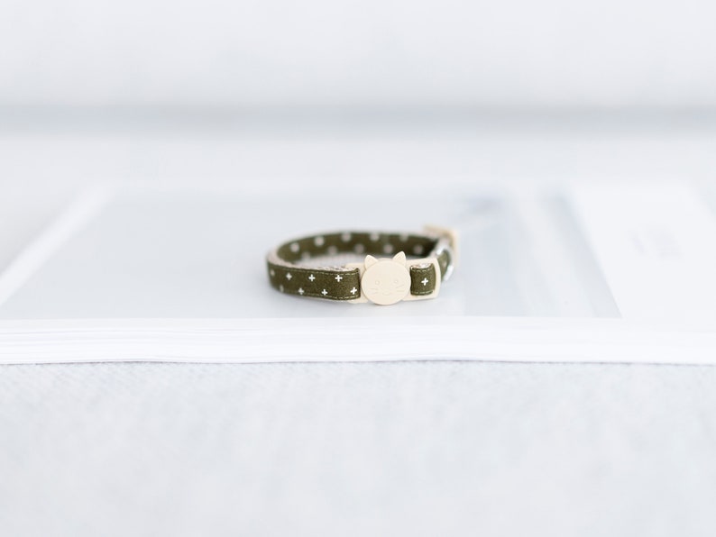 May include: Olive green cat collar with white plus signs and small white dots. The collar has a cream-colored cat-shaped buckle. A stylish accessory for your feline friend.