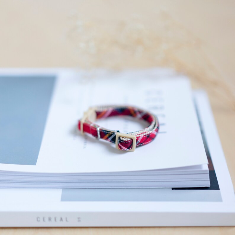May include: A red, black, and yellow plaid patterned collar with a beige buckle sits atop a stack of white paper. The collar is likely for a pet, and the design is a classic tartan pattern.