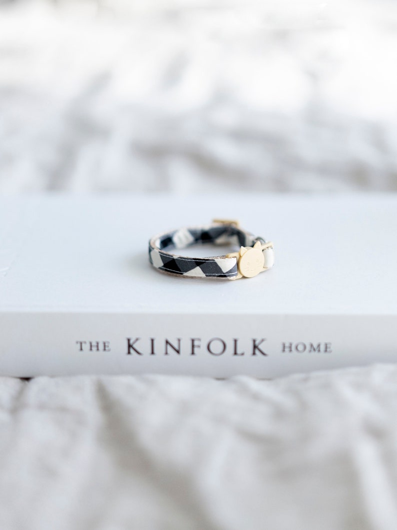 May include: A black and white patterned cat collar with a gold-colored cat-shaped charm. The collar is resting on a white book titled "THE KINFOLK HOME". The background is a soft, blurred white.