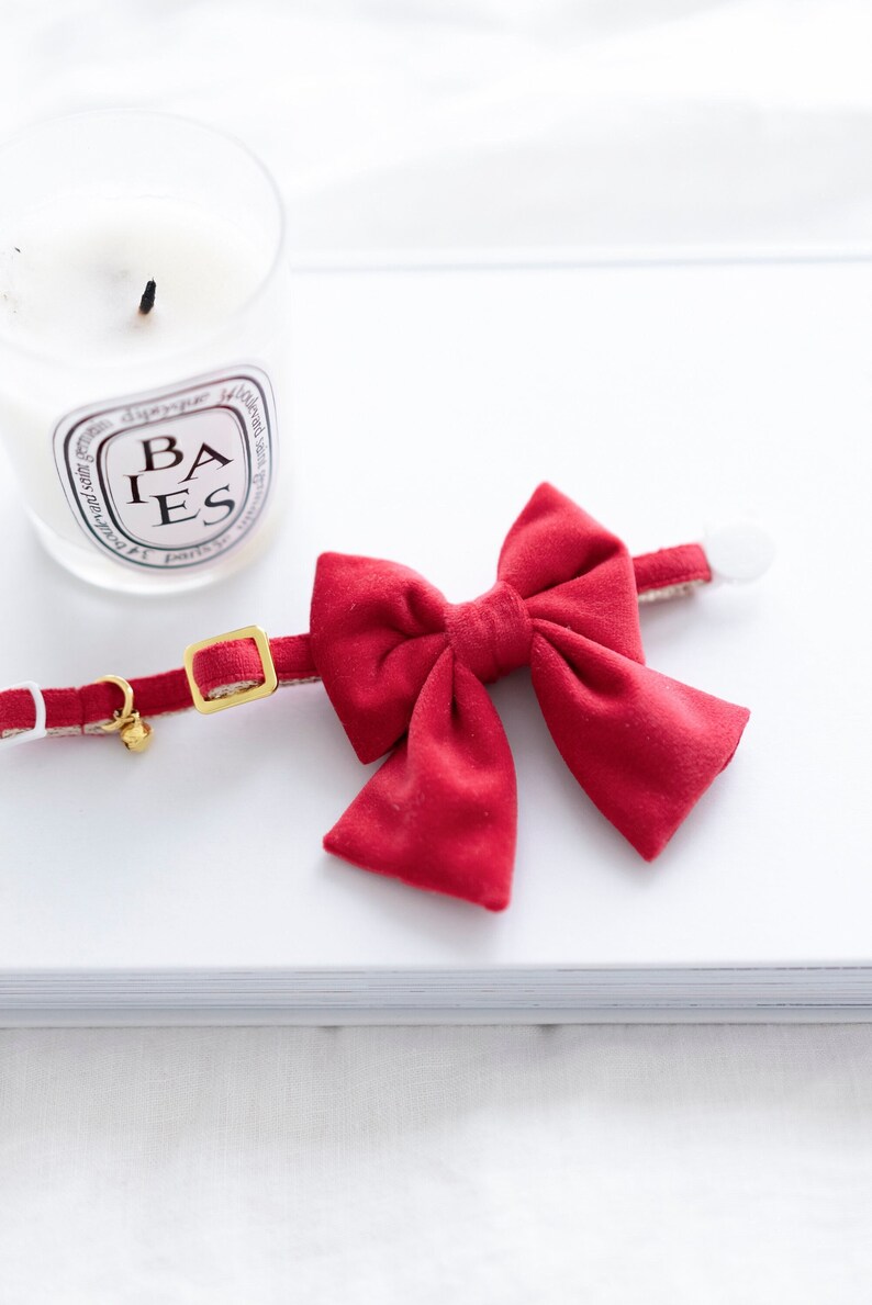 May include: A red velvet bow tie collar for pets. The collar has a gold-colored buckle and a small bell. A white candle with the text "BAIES" is in the background.