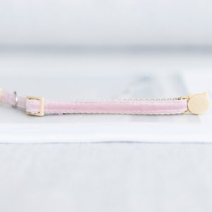 May include: A light pink velvet cat collar with a gold-colored buckle and a cat-shaped charm. The collar is a soft pink color and has a delicate, elegant appearance. The cat charm is a light beige color.