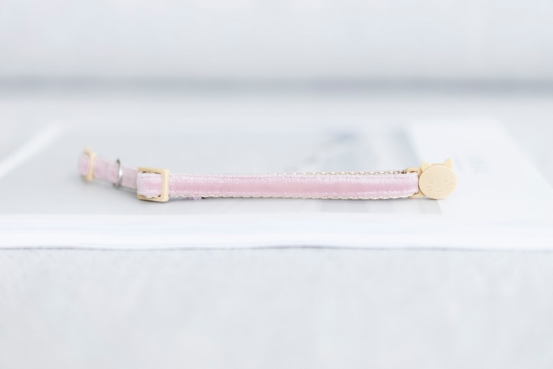 May include: A light pink velvet cat collar with a gold-colored buckle and a cat-shaped charm. The collar is a soft pink color and has a delicate, elegant appearance. The cat charm is a light beige color.