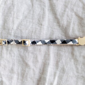 May include: A cat collar with a black and white checkered pattern. The collar has a beige plastic buckle and a cat-shaped charm. The collar is approximately 12 inches long.