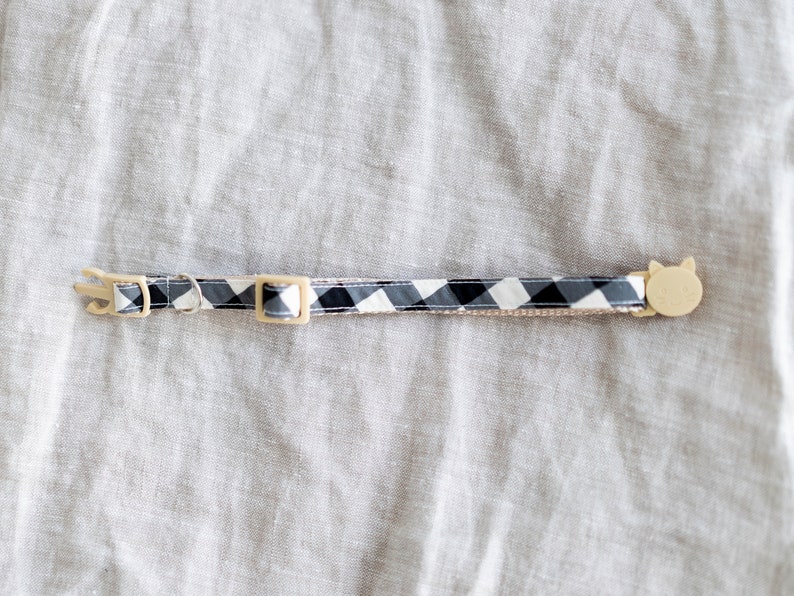 May include: A cat collar with a black and white checkered pattern. The collar has a beige plastic buckle and a cat-shaped charm. The collar is approximately 12 inches long.