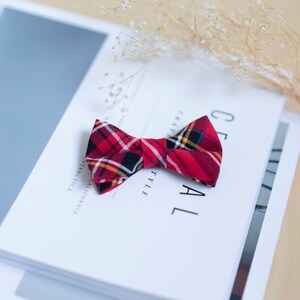 May include: A red plaid bow tie with black, yellow, and white accents rests on a stack of white books. The bow tie is made of fabric and has a classic shape. The books have text that reads "CEREAL STYLE" and "ANTARCTICA CALIFORNIA".