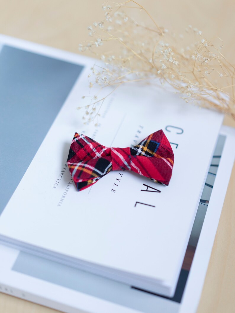 May include: A red plaid bow tie with black, yellow, and white accents rests on a stack of white books. The bow tie is made of fabric and has a classic shape. The books have text that reads "CEREAL STYLE" and "ANTARCTICA CALIFORNIA".