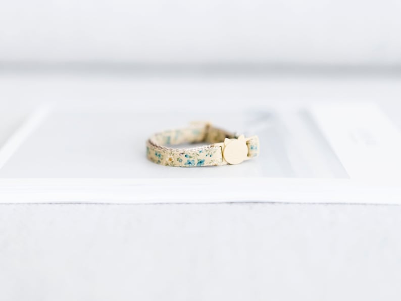 May include: A beige cat collar with a floral pattern of small blue flowers. The collar has a cream-colored cat-shaped buckle. The collar is approximately 0.5 inches wide.