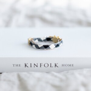 May include: A black and white checkered cat collar with a gold-colored buckle. The collar is resting on a white book titled "THE KINFOLK HOME". The background is a soft, neutral color.