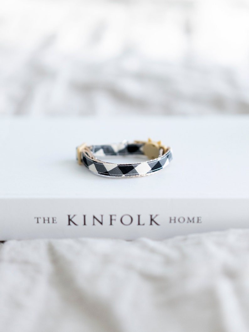 May include: A black and white checkered cat collar with a gold-colored buckle. The collar is resting on a white book titled "THE KINFOLK HOME". The background is a soft, neutral color.