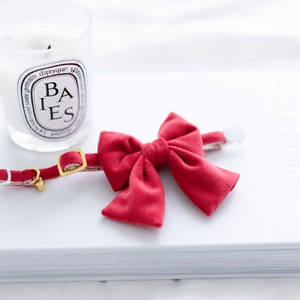May include: A red cat collar with a matching bow tie. The collar has a gold-colored buckle and a small bell. A lit candle in a glass jar is in the background.