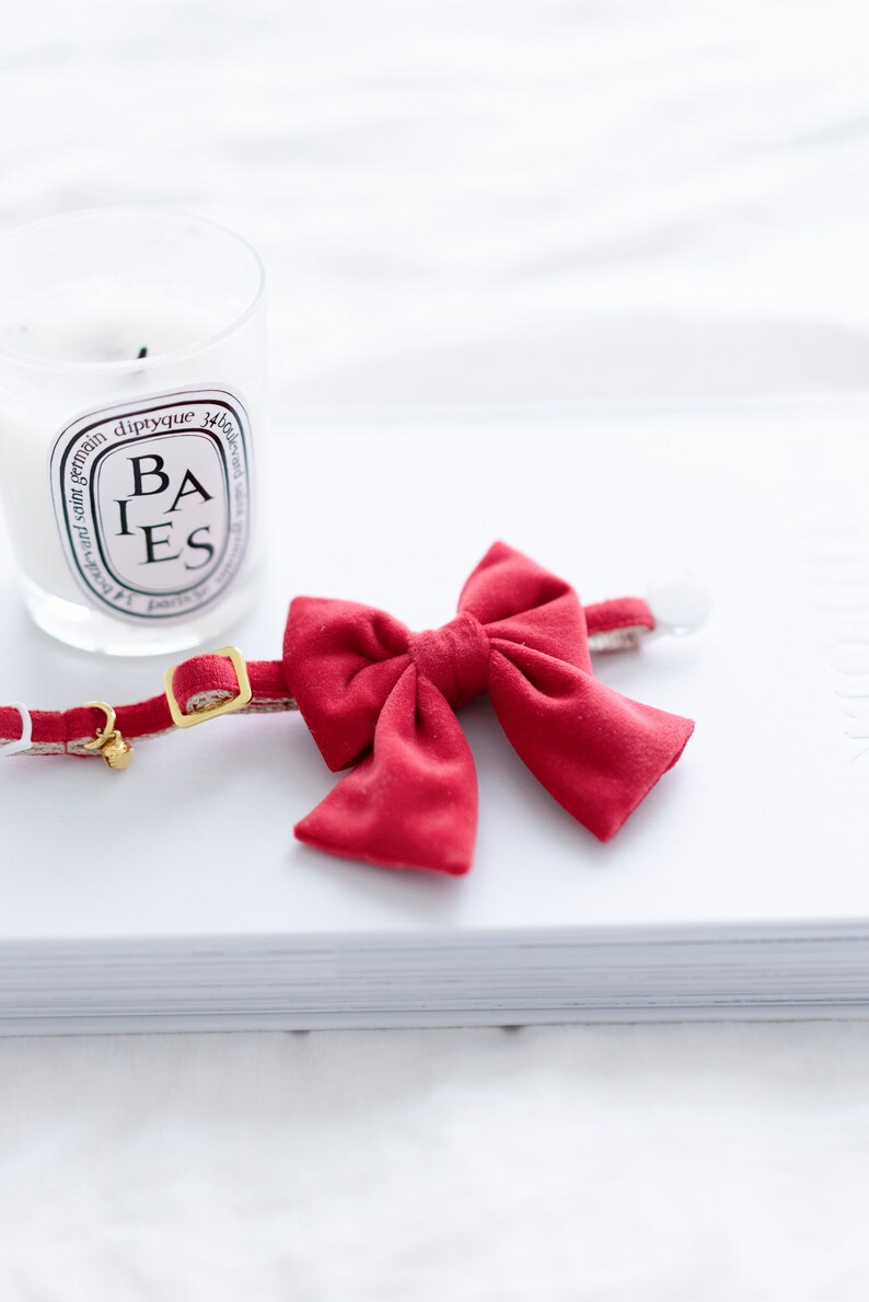 May include: A red cat collar with a matching bow tie. The collar has a gold-colored buckle and a small bell. A lit candle in a glass jar is in the background.