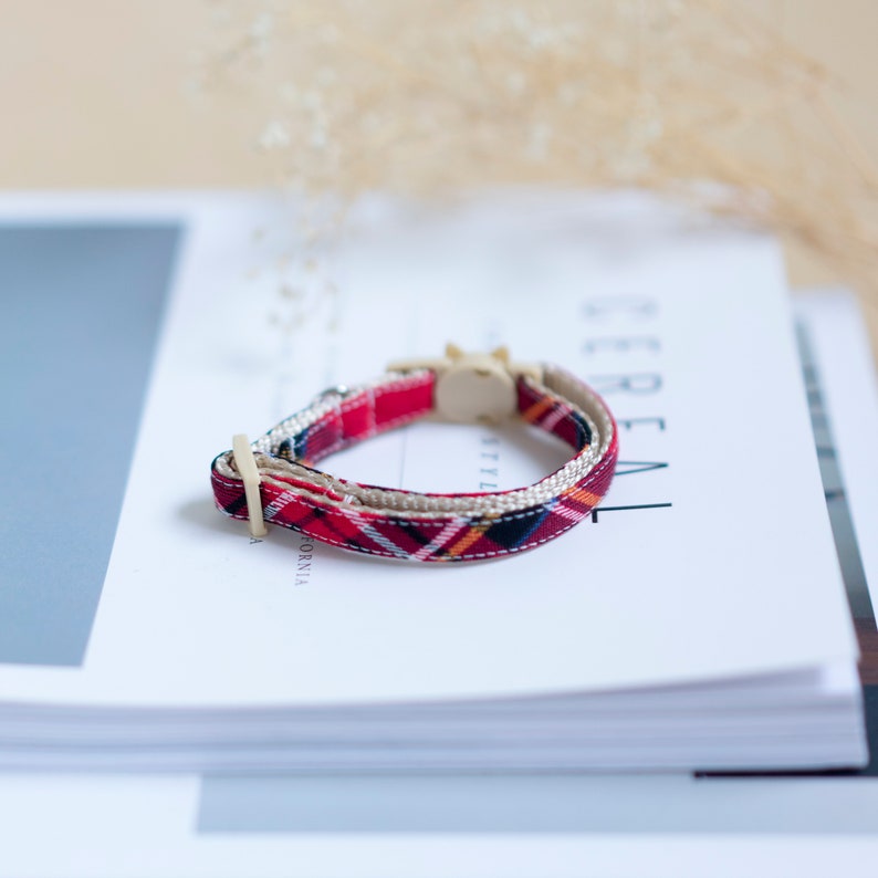 May include: A red plaid cat collar with a beige inner lining and a cream-colored buckle. The collar features a classic plaid pattern in red, black, yellow, and white. The collar is approximately 10 inches in length.