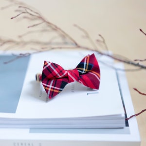 May include: A red plaid bow tie with black, white, and yellow accents. The bow tie is made of fabric and is sitting on a stack of white books. The bow tie is a classic accessory for formal events.