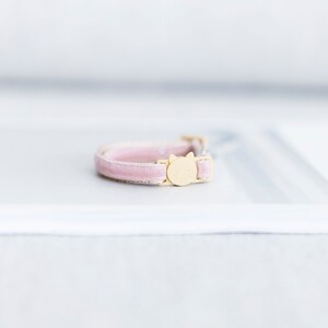 May include: A pink velvet cat collar with a gold-colored buckle featuring a cat face design. The collar is a soft pink color and appears to be made of a plush material. The buckle is a light gold color.