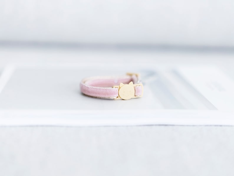 May include: A pink velvet cat collar with a gold-colored buckle featuring a cat face design. The collar is a soft pink color and appears to be made of a plush material. The buckle is a light gold color.
