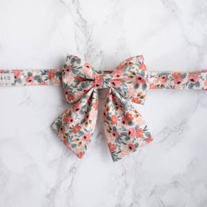 May include: A dog collar and bow tie set with a floral design. The cream-coloured fabric is patterned with pink roses, grey leaves, and blue accents. The bow tie is large and the focal point of the image.