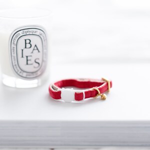 May include: A red cat collar with a white cat-shaped buckle and a small gold bell. The collar is made of a soft, velvety material. A white candle with black text is in the background.
