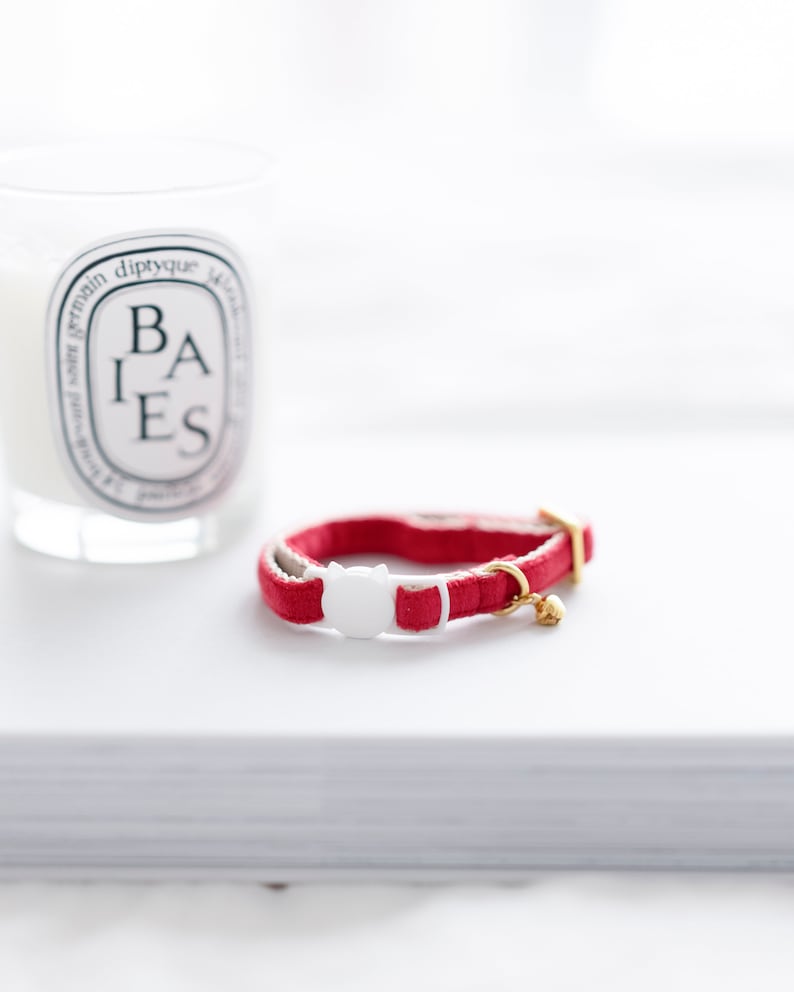 May include: A red cat collar with a white cat-shaped buckle and a small gold bell. The collar is made of a soft, velvety material. A white candle with black text is in the background.