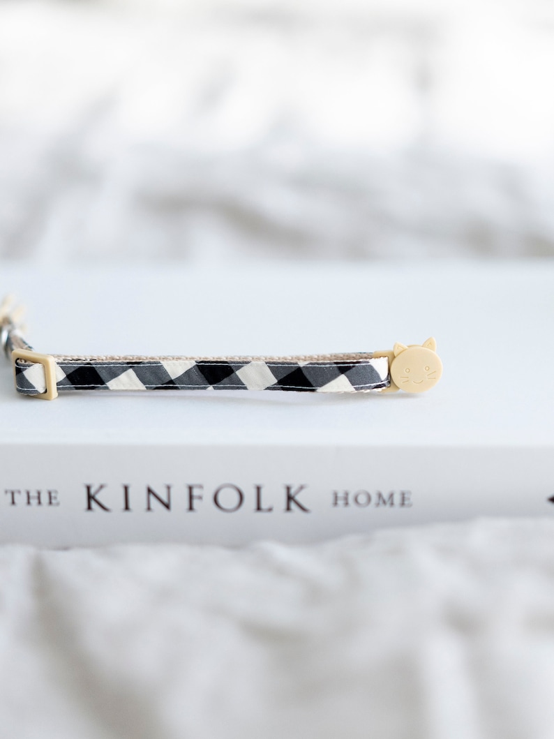 May include: A black and white checkered cat collar with a beige cat-shaped buckle. The collar is resting on a white book with the text "THE KINFOLK HOME". The collar is a pet accessory.