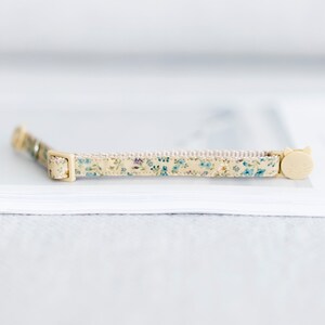 May include: A beige cat collar with a floral pattern of blue and green flowers. The collar has a cream-colored buckle and a cat-shaped clasp. The collar is approximately 12 inches long.