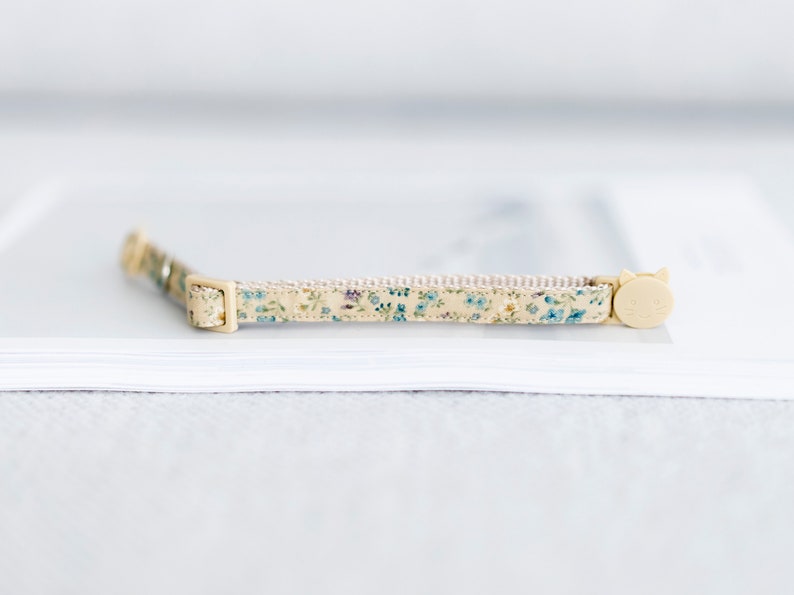 May include: A beige cat collar with a floral pattern of blue and green flowers. The collar has a cream-colored buckle and a cat-shaped clasp. The collar is approximately 12 inches long.