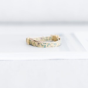 May include: A beige floral cat collar with blue and purple flowers. The collar has a cream-colored buckle and a woven trim. The collar is approximately 10 inches long.