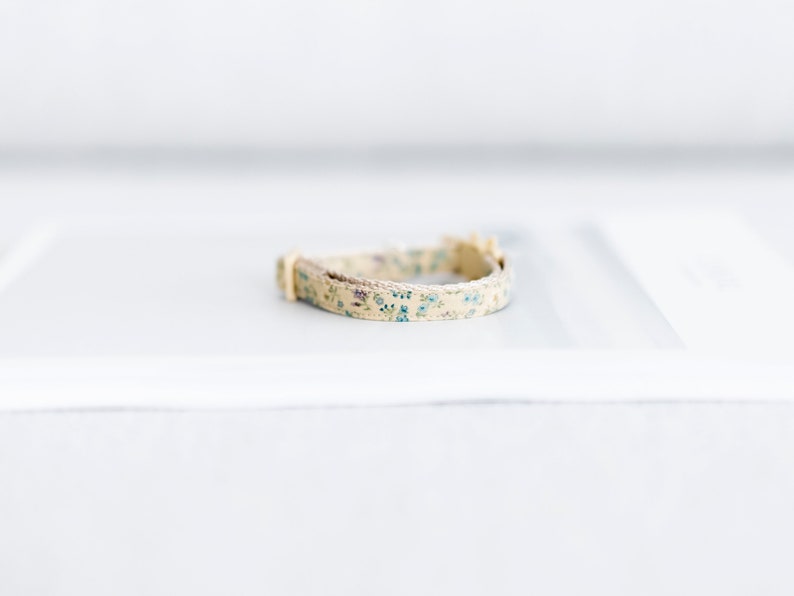 May include: A beige floral cat collar with blue and purple flowers. The collar has a cream-colored buckle and a woven trim. The collar is approximately 10 inches long.