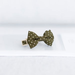 May include: Olive green bow tie with a white cross pattern, attached to a beige collar with a square buckle. The bow tie is centered on a white surface, ideal for pets.
