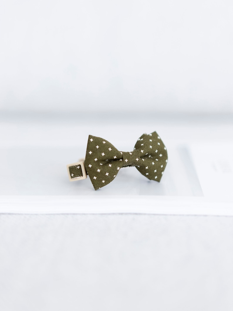 May include: Olive green bow tie with a white cross pattern, attached to a beige collar with a square buckle. The bow tie is centered on a white surface, ideal for pets.