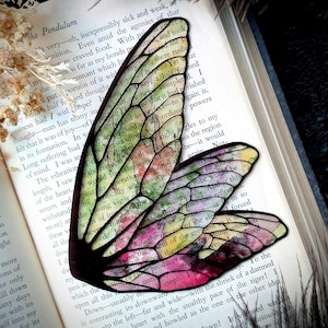 May include: A colorful, translucent fairy wing, with black outlines and a watercolor effect, rests on an open book. The wing features shades of pink, green, yellow, and purple. The book's pages display printed text.