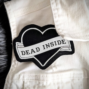May include: Black heart-shaped patch with a white banner that reads "DEAD INSIDE" on a white denim jacket.