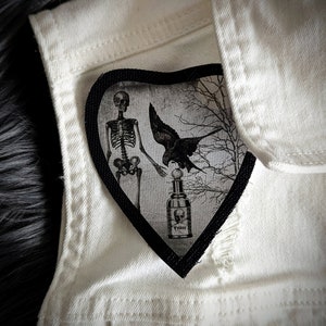 May include: A heart-shaped patch with a black border featuring a skeleton, raven, and poison bottle design. The patch is set against a white denim jacket, creating a gothic aesthetic. The bottle has the word "POISON" on it.