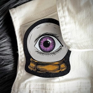 May include: A black and white illustration of a crystal ball with a purple eye inside. The crystal ball is on a gold stand.
