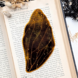 May include: A close-up of a brown and gold butterfly-shaped bookmark with a pattern of stars. The bookmark is placed on an open book with the text "Pit and the Pendulum" visible.