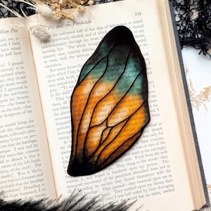 May include: A close-up of a stained glass butterfly bookmark with orange, yellow, green, and blue colors. The bookmark is resting on a page of a book titled "Pit and the Pendulum" by Edgar Allan Poe.