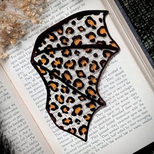 May include: A decorative bookmark with a leopard print design. The bookmark is shaped like a bat with black outlines and brown and orange spots. It is placed on top of an open book.