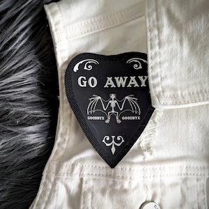 May include: Black heart-shaped patch with the text "GO AWAY" in white lettering. A skeleton bat with the text "GOODBYE" on either side of the bat is below the text. The patch is on a white denim jacket.