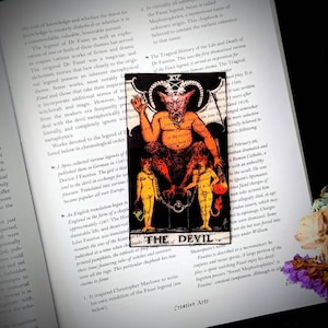 May include: The Devil tarot card from the Rider-Waite deck. The card features a red, horned, and winged demon with a human-like body. The demon is sitting on a throne with a chain and a key. Two naked figures are chained to the throne. The card is set against a black background with gold accents. The text "THE DEVIL" is printed in gold at the bottom of the card.
