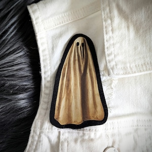 May include: A brown and white ghost-shaped iron-on patch with a black border. The patch is on a white denim jacket.